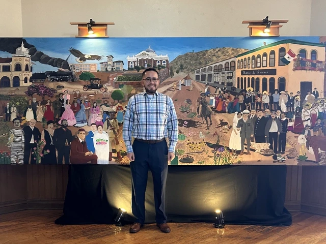 Man wearing a plaid shirt, dark pants, and glasses stand in front of a mural showing the past of Santa Cruz County