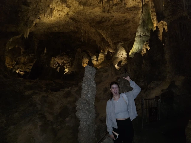 A young women wearing black pants and gray sweater is in a cavern and pointing up to the ceiling