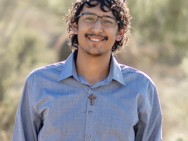 Young man with curly black hair and wearing glasses and a blue shirt, stands outside smiling
