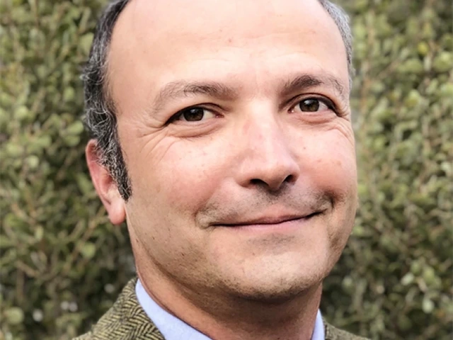 Man in a blue shirt and brown sports coat stand against a hedge, smiling