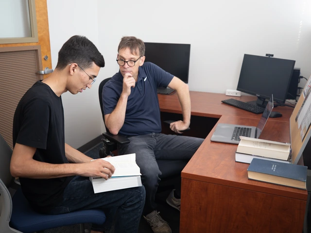 A professor and student sit together at a desk while the student holds a book and they are talking