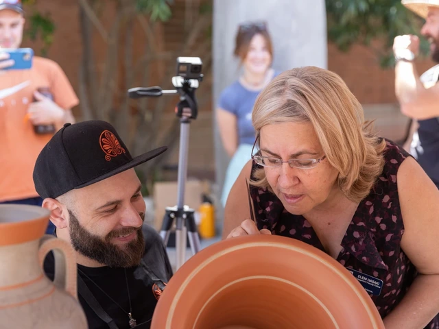 A woman with blonde hair and glasses bends over a piece of pottery to look closely and a man with a black hat and a beard sits near her, looking at the same piece
