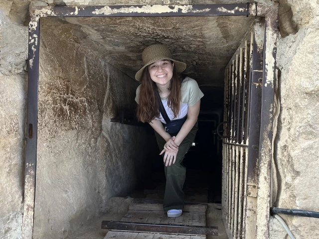 A woman with long brown hair and wearing a hat crouches down, emerging from a mining shaft