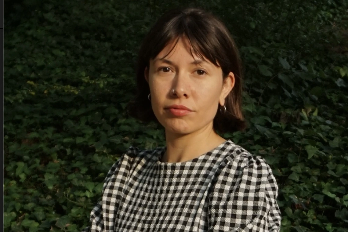 A woman with dark brown hair and wearing a black and white checked dress, stands outside with arms folded