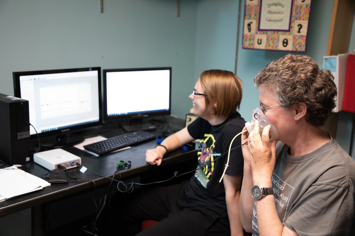 A professor and student sit in front of computer screens while using a breathing apparatus tool for research
