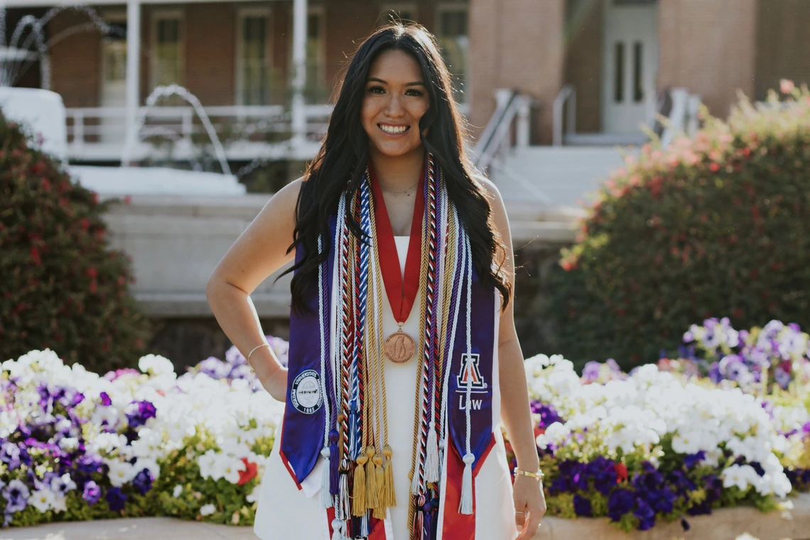 Student with long brown hair stands in front of Old Main, wearing graduation regalia