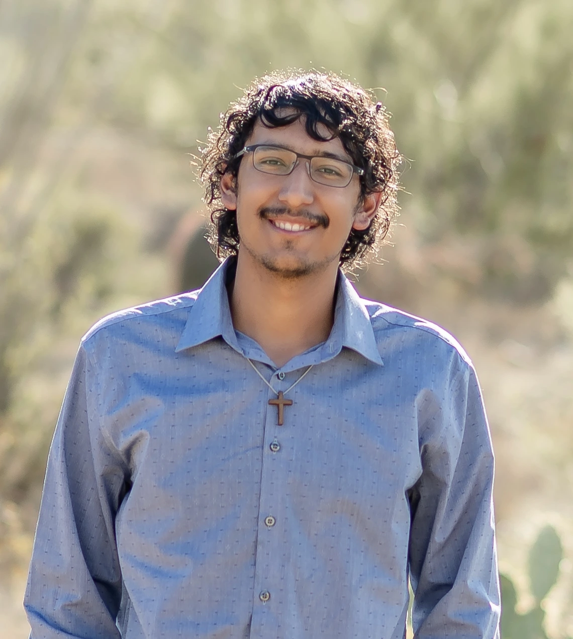 Young man with curly black hair and wearing glasses and a blue shirt, stands outside smiling