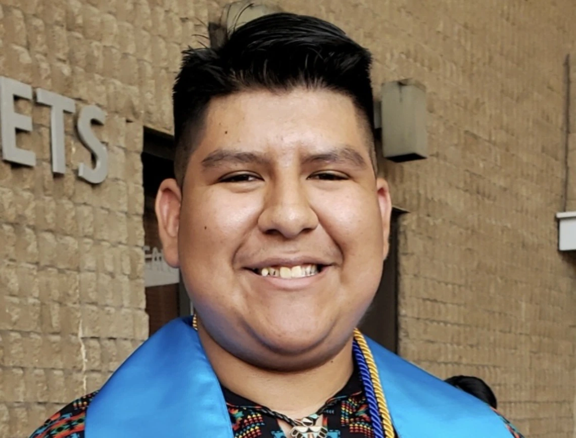 Student stands outside a brick building, wearing graduation regalia