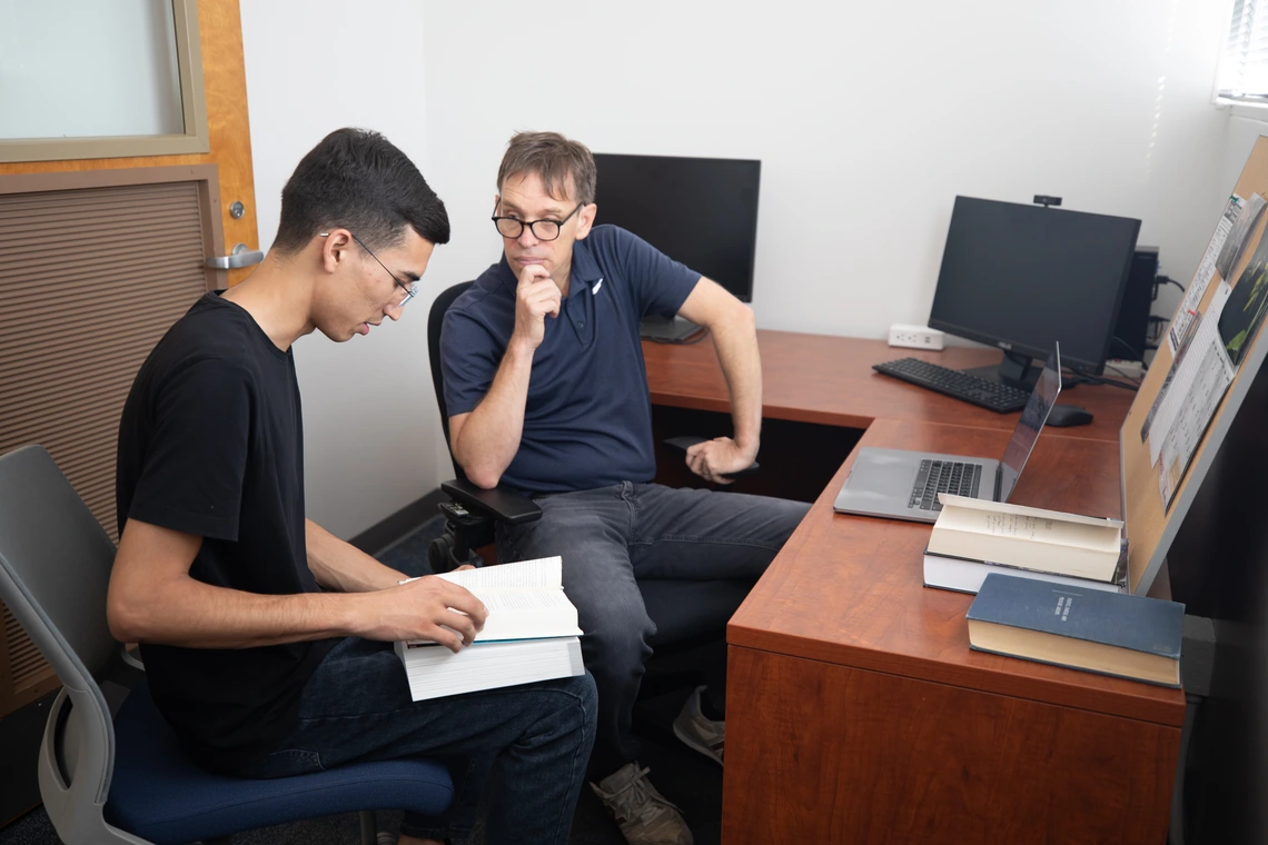 A professor and student sit together at a desk while the student holds a book and they are talking