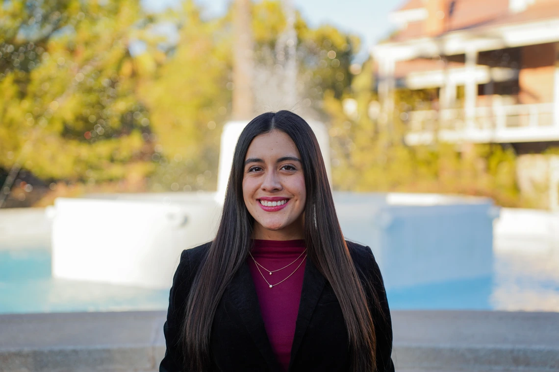 Young woman with long dark hair wearing a black jacket and maroon shirt, stands outside in front of a fountain