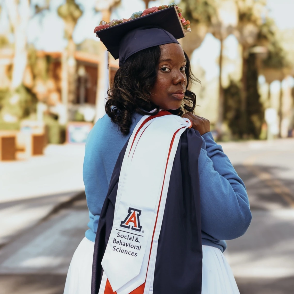 A graduating student stands outdoors wearing a navy cap decorated with flowers and a white graduation stole with the University of Arizona logo and “Social & Behavioral Sciences” printed on it. She looks back over her shoulder toward the camera, with trees and campus buildings blurred in the background.