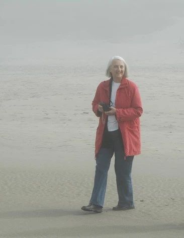 Woman with bobbed gray hair and wearing a red jacket stands on a beach