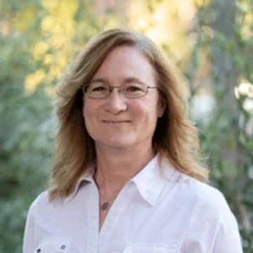 WOman with blonder hair, glasses and wearing a white shirt stands outside