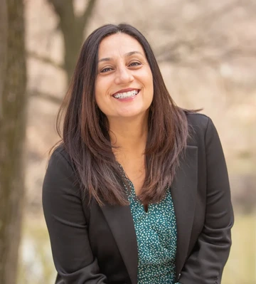 Woman with long brown hair and wearing a green top and black jacket stands outside smiling