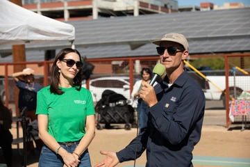 A woman on the left in a green Sprouts t-shirt looks to her left at a man in a blue shirt and baseball cap, who holds a microphone and is speaking