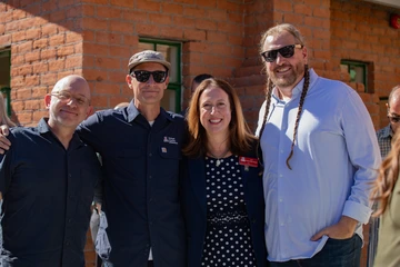 Three men and one woman stands together in a row, outside, in front of a brick home