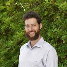 Man with dark brown hair, beard and wearing a white shirt stands in front of greenery outside