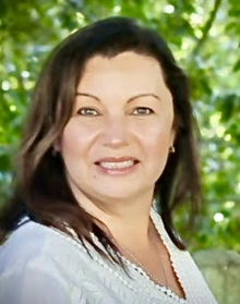 Woman with brown hair and standing outside in a treed setting, smiles for the camera