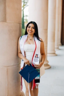Woman stands by a pillar outside and wears white graduation regalia