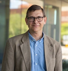 Man with brown hair and glasses, wearing blue shirt and brown jacket