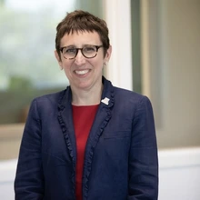 A woman with short brown hair and glasses stands outside wearing a red jacket and blue shirt