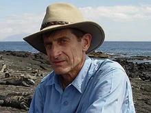 Man in a blue shirt and wearing a brown wide brimmed hat sits with the ocean at his back