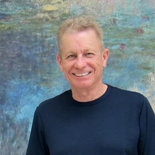 Man with blonde hair and wearing a black shirt stands in front of a watercolor painting