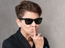 Woman with short hair, wearing black Ray Ban like sunglasses and her pointer finger resting on her lips