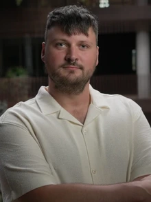 A student with short dark hair and a trimmed beard, wearing a light-colored short-sleeve shirt, stands with their arms crossed in an indoor setting with soft lighting and blurred architectural elements in the background