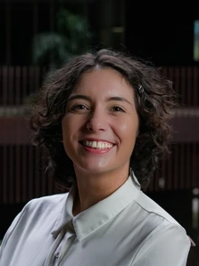 A student with short, curly dark hair smiles at the camera. They are wearing a light-colored collared blouse and is photographed indoors against a softly blurred background